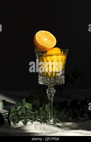Ancora vita giallo limone intero e mezzo in bicchiere di vino e ramo di plastica con foglie verdi su sfondo nero alla luce del sole Foto Stock