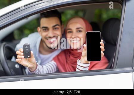 Felice moglie islamica in hijab e marito mostra chiave e smartphone con schermo vuoto in finestra aperta in auto Foto Stock