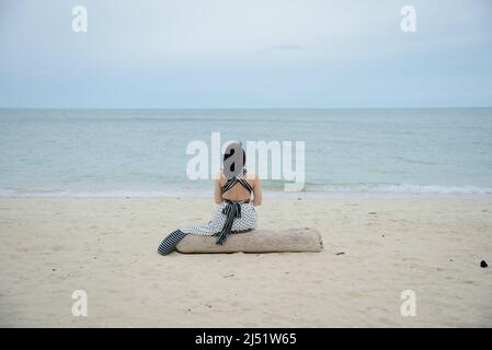 Vista posteriore di una donna solitaria seduta sul tronco e guardando al mare. Foto Stock
