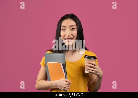 Felice studentessa coreana con taccuini e caffè da asporto, in piedi su sfondo viola Foto Stock