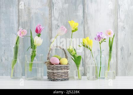 Fiori colorati in bottiglie di vetro e uova di Pasqua in cesto di vimini sul tavolo. Sfondo messaggi di saluto, spazio di copia. Foto Stock