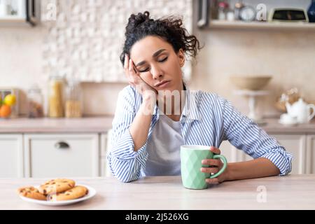 Giovane donna addormentata seduta al tavolo da pranzo in cucina con gli occhi chiusi, tenendo una tazza mentre si fa colazione Foto Stock