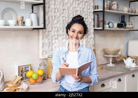Felice donna latina tenendo libro di cucina, scrittura ricetta per cena deliziosa, in piedi in cucina e sorridendo alla macchina fotografica Foto Stock