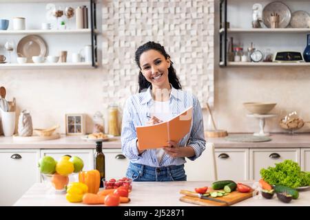 Concetto di alimentazione e dieta sana. Donna eccitata che fa la lista del cibo necessario alla cucina, tenendo il taccuino con la penna Foto Stock