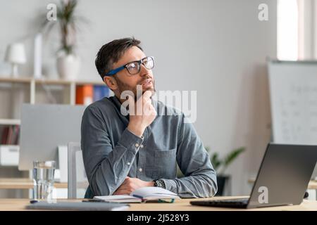 Uomo maturo e premuroso seduto in ufficio e guardando da parte, lavorando con il notebook, alla ricerca di idee creative Foto Stock