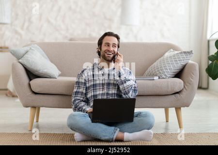 Felice uomo europeo millenario con le chiappe in camicia telefona, lavora su un notebook, si siede sul pavimento in salotto Foto Stock