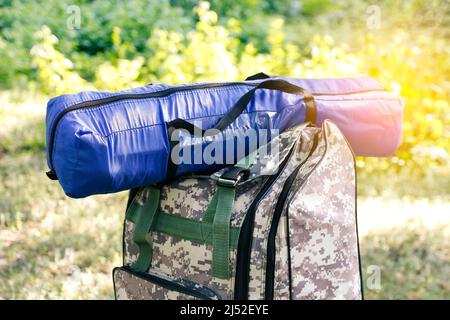 Zaino militare defocus e tenda blu o sacco a pelo. Borsa esercito su sfondo verde erba vicino albero. Zaino militare mimetizzazione. Turistico Foto Stock