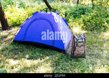 Sfocatura blu tenda turistica aperta in piedi su sfondo verde natura. Zaino militare. Concetto di turismo. Vacanze estive nella foresta, campeggio. Fuori fuoco Foto Stock