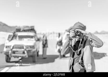 Un uomo irriconoscibile e defocused con il foulard berbero che fotografa con una fotocamera DSLR nella strada del deserto del Sahara con una macchina 4x4 sfocata e uomini tuareg. Foto Stock