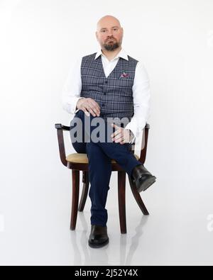 Ritratto di splendido uomo d'affari di mezza età in gilet grigio, jeans blu, camicia bianca seduta su sedia con gambe incrociate. Foto Stock