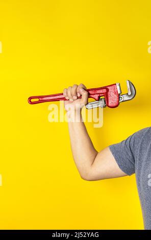 Braccio dell'uomo irriconoscibile che tiene una chiave a tubo con il suo muscolo flettente del braccio su uno sfondo giallo. Concetto di giorno di lavoro Foto Stock