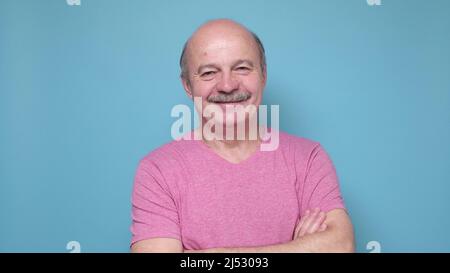 Bell'uomo anziano che mantiene le braccia incrociate e sorridenti mentre si alza isolato su sfondo blu. Foto Stock