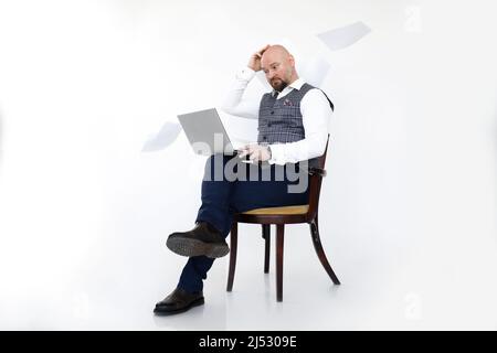 Ritratto di uomo di mezza età perplesso in gilet grigio, jeans blu, camicia bianca, seduta con laptop tra fogli di carta. Foto Stock