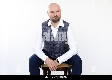 Ritratto di uomo d'affari di mezza età in gilet grigio, jeans blu, camicia bianca seduta su sgabello di legno su sfondo bianco. Foto Stock