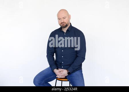 Ritratto di uomo di mezza età in camicia blu, jeans seduti su sgabello, dita interlacciate su sfondo bianco. Foto Stock