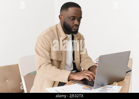 Focalizzato giovane afro americano uomo contando le spese e mantenendo i record su laptop, conti bancari cartacei sono sul tavolo. Il modello maschile gestisce hous mensile Foto Stock