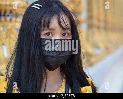 Attraente ragazza thailandese con gli occhi scuri belli e lunghi capelli neri indossa una maschera protettiva monouso nera di fronte ad un tempio buddista wat. Foto Stock