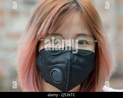 Giovane donna vietnamita graziosa con i capelli tinti e gli occhiali indossa una maschera protettiva nera con valvola di respirazione durante la pandemia di coronavirus. Foto Stock