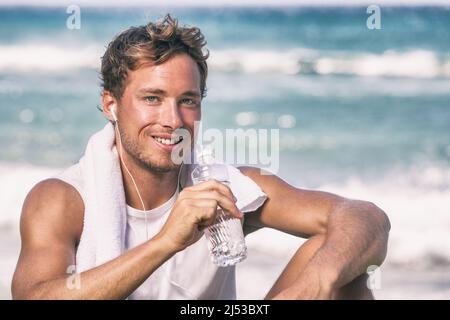 Atleta giovane in salute che beve acqua bottiglia dopo l'allenamento in spiaggia ascoltando musica app telefono con auricolari. Ritratto sorridente Foto Stock