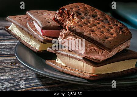 Sandwich al gelato su un piatto nero con un tovagliolo blu sul fondo su un tavolo di legno con sfondo nero, illuminato da una luce della finestra Foto Stock