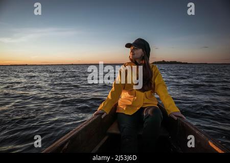 Giovane donna in un impermeabile giallo e cappellino che naviga su una barca e guarda lontano Foto Stock