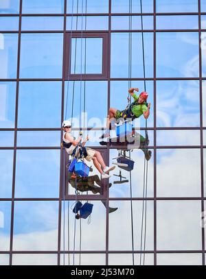 Lavoratori di alpinismo industriale pulitori professionali lavando finestre in vetro, appesi su funi di arrampicata di sicurezza. Il lavaparabrezza guarda la telecamera e mostra i bicipiti mentre il collega dà i pollici in su. Foto Stock