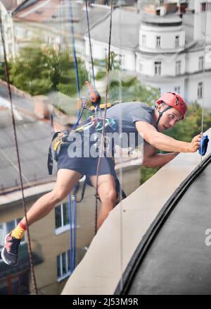 Alpinista industriale appeso su corda e finestra di pulizia con panno in microfibra. Vista dall'interno dell'edificio. Pulire utilizzando un'attrezzatura di sollevamento di sicurezza durante la pulizia del vetro di un edificio alto. Foto Stock