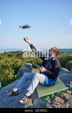 Uomo che utilizza il drone tramite telecomando. Uomo che utilizza il drone al tramonto per foto e video making mentre si siede in cima al masso alto in montagna. Foto Stock