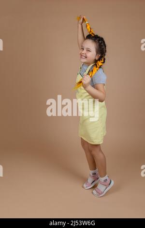 Bambina giocosa con trecce gialle kanekalon, saltando con le mani che tengono i pigtail guardando la macchina fotografica che indossa tuta gialla e t-shirt grigia Foto Stock