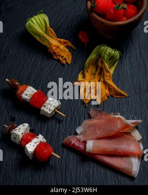 Due spiedini con pezzi di formaggio e pomodori ciliegini su un piatto di ardesia, con fiori di zucchine e prosciutto affettato accanto a loro Foto Stock