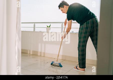 Uomo che pule il pavimento con il mop. Il maschio del janitor che lava i pavimenti sporchi. Guy mopping il balcone in una giornata di sole. Persona che fa i chores domestici. Giovane collega Foto Stock