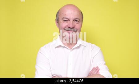 Bell'uomo anziano che mantiene le braccia incrociate e sorridenti mentre si trova isolato su sfondo giallo. Foto Stock