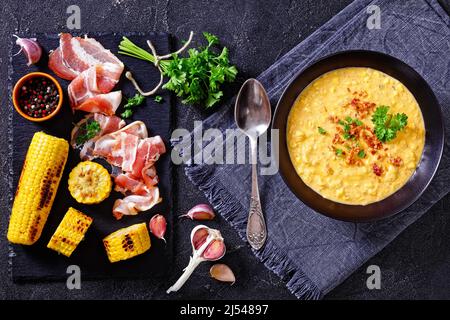 zuppa di mais con pancetta fritta croccante sbriciolata in ciotola nera su tavola di cemento con ingredienti, vista orizzontale dall'alto, posa piatta Foto Stock