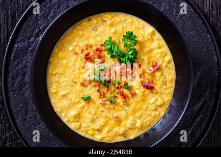 zuppa di mais con pancetta fritta croccante sbriciolata in una ciotola nera su un tavolo di legno scuro, vista orizzontale dall'alto, posa piatta, primo piano Foto Stock