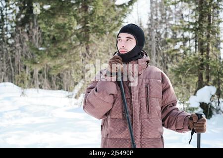 Ritratto di giovane uomo caucasico che indossa balaclava trekking o sci nella foresta locale in soleggiata giornata invernale guardando lontano Foto Stock