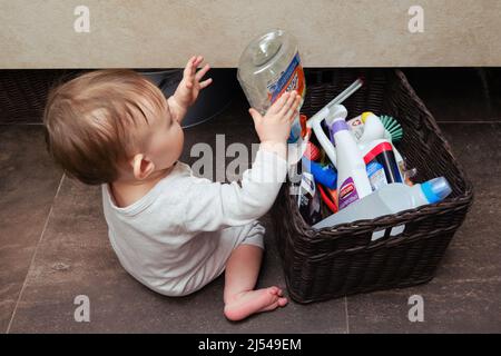 Il bambino del bambino del toddler sta giocando nella stanza del gabinetto con i detersivi di pulizia del muscolo del sig... Il bambino gioca su un pavimento marrone in un bagno beige con il ch della casa Foto Stock