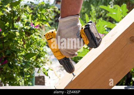 Un falegname lavora con un cacciavite elettrico giallo, ruota una vite in un asse di legno trave Foto Stock