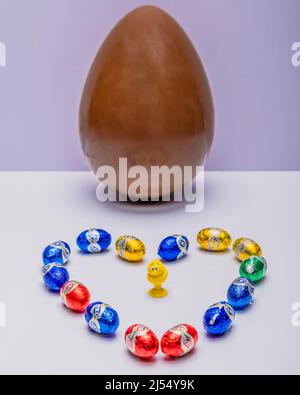 Piccole uova di cioccolato colorate a forma di cuore con un uovo di Pasqua sullo sfondo Foto Stock