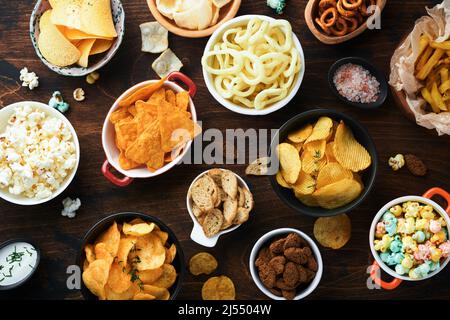 Cibo o spuntini malsani. Tutti i classici spuntini di patate con arachidi, anelli di popcorn e cipolle e pretzel salati in piatti da ciotola su vecchio sfondo di legno. Foto Stock