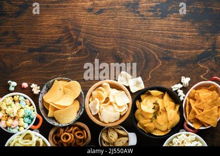 Cibo o spuntini malsani. Tutti i classici spuntini di patate con arachidi, anelli di popcorn e cipolle e pretzel salati in piatti da ciotola su vecchio sfondo di legno. Foto Stock