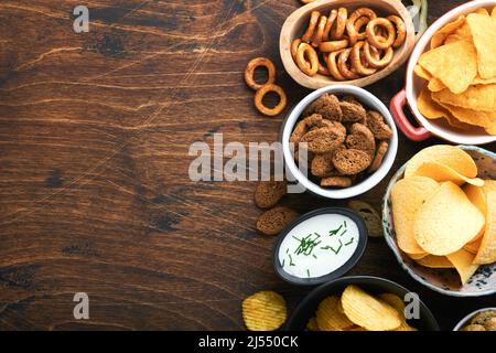 Cibo o spuntini malsani. Tutti i classici spuntini di patate con arachidi, anelli di popcorn e cipolle e pretzel salati in piatti da ciotola su vecchio sfondo di legno. Foto Stock