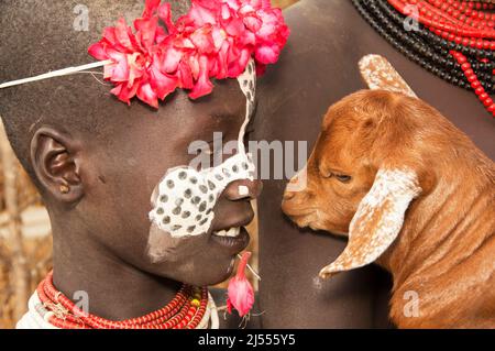 Karo ragazza con una fascia floreale, dipinti facciali, collane colorate, piercing labbro e una capra nelle braccia, Omo valle del fiume, Etiopia meridionale Foto Stock