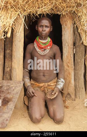 Donna Karo con un sacco di collane colorate e conchiglie di cowry, piercing labbro e dipinti facciali di fronte alla sua capanna, valle del fiume Omo, Sud Etiopia Foto Stock