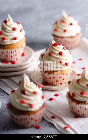 Muffin alla vaniglia con glassa alla crema spolverata con cuori di zucchero rosso sui piatti e un panno bianco, su sfondo grigio. Foto Stock