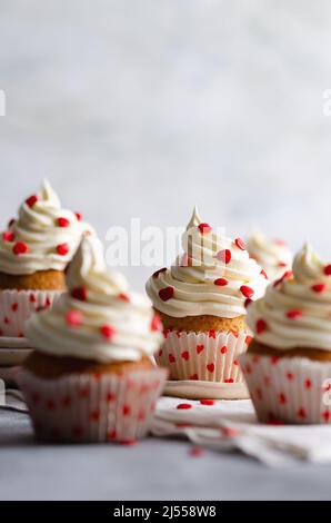 Muffin alla vaniglia con glassa alla crema spolverata con cuori di zucchero rosso sui piatti e un panno bianco, su sfondo grigio con spazio per la copia. Foto Stock