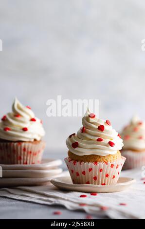 Muffin alla vaniglia con glassa alla crema spolverata con cuori di zucchero rosso sui piatti e un panno bianco, su sfondo grigio con spazio per la copia. Foto Stock