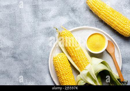 Tre pannocchie di mais, una ciotola con farina di mais e un cucchiaio di legno in un piatto su sfondo grigio. Foto Stock