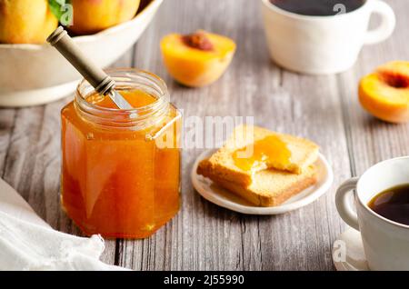 Marmellata di pesca fatta in casa in un barattolo con alcune pesche fresche, due tazze di caffè e alcuni toast, su uno sfondo di legno. Foto Stock