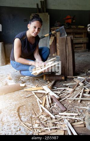 fotografia verticale di una donna sorridente accovacciata a terra raccogliendo legna da ardere tagliata di fresco per fare un fuoco. uguaglianza sul lavoro. empowerment. forza. Foto Stock