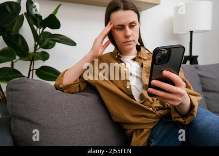 Fuoco selettivo di donna infelice pensierosa che guarda lo schermo del cellulare, in attesa di chiamata dal ragazzo. Messaggi di lettura femminili o e-mail con cattivo Foto Stock